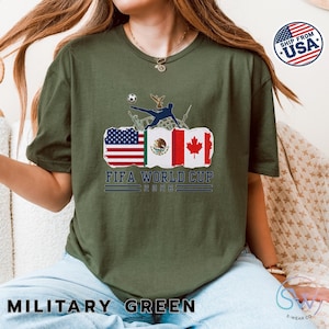 May include: Military green t-shirt featuring a graphic for the FIFA World Cup 2026. The design includes flags of the USA, Mexico, and Canada, with a soccer player kicking a ball. The text "FIFA WORLD CUP 2026" is displayed below the flags.