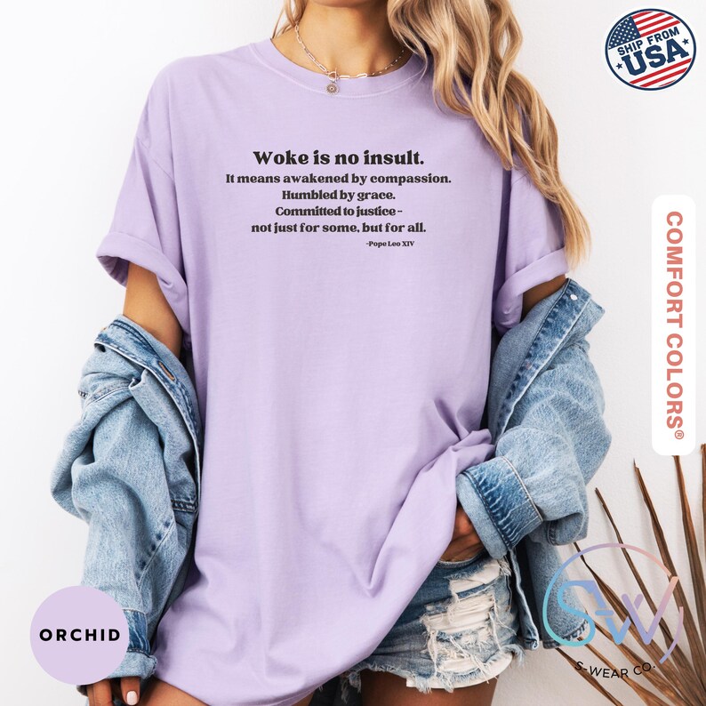 May include: A light purple t-shirt with the text "Woke is no insult" and a quote by Pope Leo XIV. The shirt is part of the Comfort Colors collection and is made in the USA. The model is wearing a denim jacket.