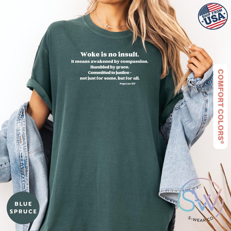 May include: Blue spruce Comfort Colors t-shirt with the text "Woke is no insult." in white. The text continues with a quote by Pope Leo XIV. The shirt has rolled-up sleeves and is paired with a denim jacket.