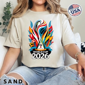 May include: A sand-colored t-shirt featuring a vibrant, multi-colored graphic of the World Cup 2026 trophy. The design includes abstract shapes in red, blue, yellow, and black, with the text World Cup 2026 printed in bold black lettering below.