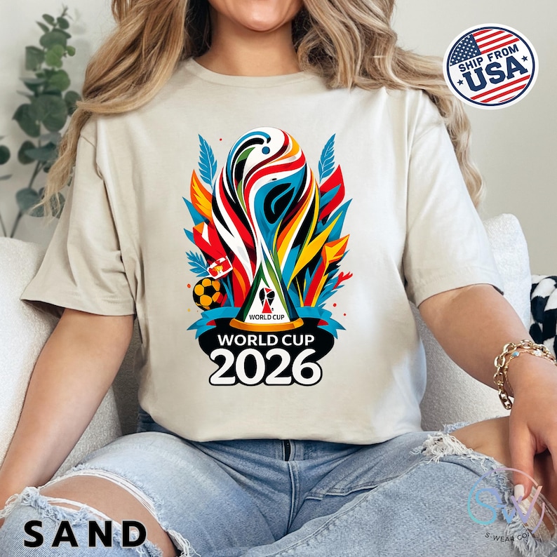 May include: A sand-colored t-shirt featuring a vibrant, multi-colored graphic of the World Cup 2026 trophy. The design includes abstract shapes in red, blue, yellow, and black, with the text World Cup 2026 printed in bold black lettering below.