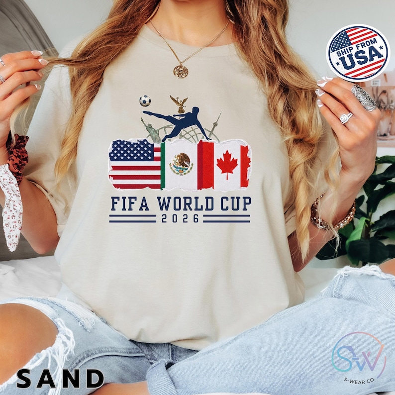 May include: Sand-colored t-shirt featuring a FIFA World Cup 2026 graphic. The design includes flags of the USA, Mexico, and Canada, along with a soccer ball and a player kicking the ball. The text "FIFA WORLD CUP 2026" is printed below the flags.