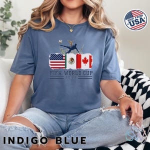 May include: Indigo blue t-shirt with a graphic featuring flags of the USA, Mexico, and Canada, a soccer player kicking a ball, and the text "FIFA WORLD CUP 2026". The shirt is a short-sleeved, crew neck style.