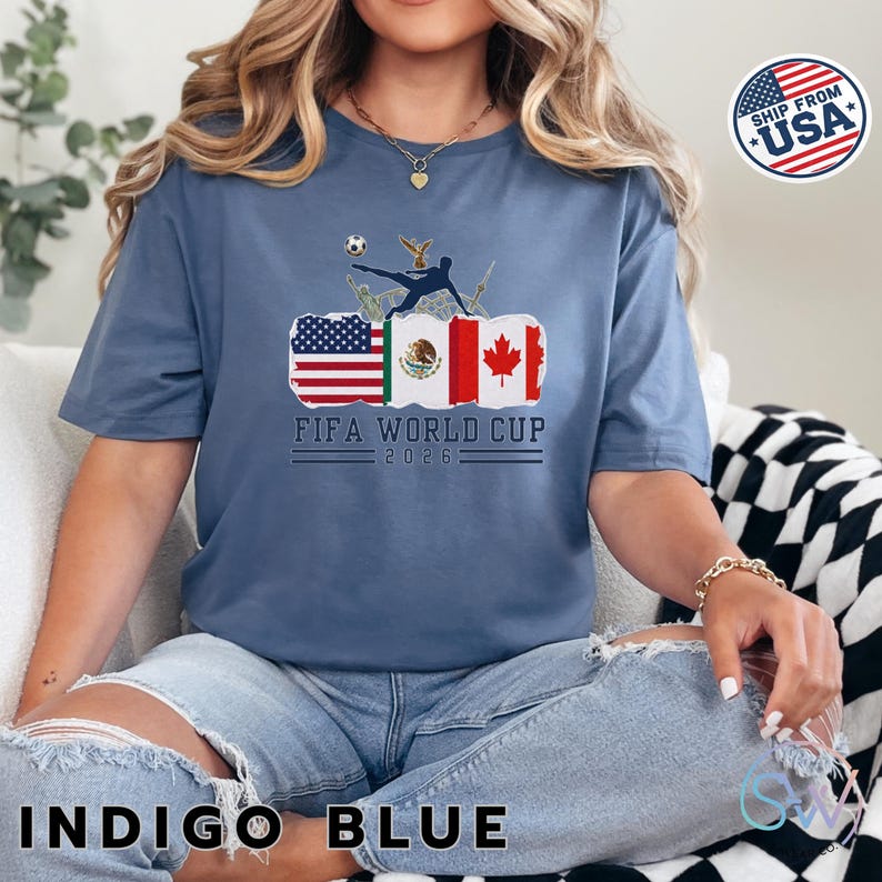 May include: Indigo blue t-shirt with a graphic featuring flags of the USA, Mexico, and Canada, a soccer player kicking a ball, and the text "FIFA WORLD CUP 2026". The shirt is a short-sleeved, crew neck style.