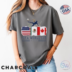 May include: Charcoal gray t-shirt featuring a graphic for the FIFA World Cup 2026. The design includes flags of the USA, Mexico, and Canada, a soccer player kicking a ball, and the text "FIFA WORLD CUP 2026".