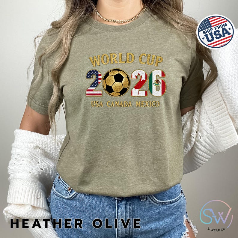 May include: Heather olive t-shirt with "WORLD CUP 2026" in gold glitter lettering. The numbers 2 and 6 are designed with the USA and Mexican flags, with a soccer ball in the center. The text "USA CANADA MEXICO" is below the numbers.