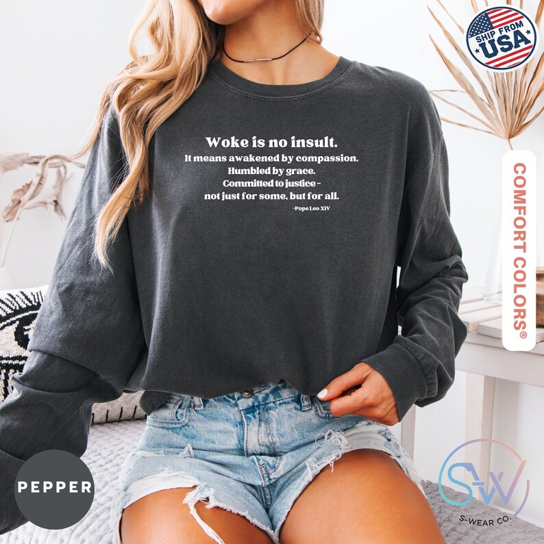May include: Dark gray long-sleeve sweatshirt with the text "Woke is no insult." in white. The text includes a quote by Pope Leo XIV. The sweatshirt is made of Comfort Colors material.