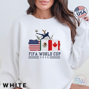 May include: White sweatshirt featuring a FIFA World Cup 2026 design. The graphic includes flags of the USA, Mexico, and Canada, a soccer player kicking a ball, and the text "FIFA WORLD CUP 2026".