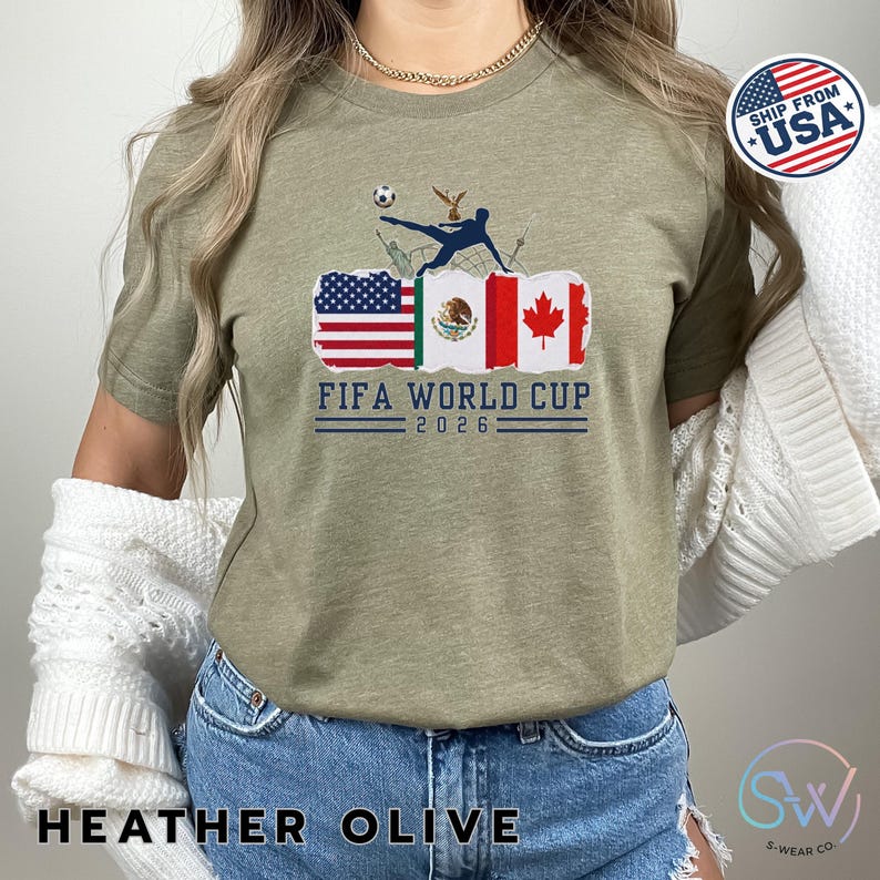 May include: Heather olive green t-shirt featuring the text "FIFA WORLD CUP 2026" with flags of the USA, Mexico, and Canada. A soccer player kicks a ball above the flags. The shirt also has the text "HEATHER OLIVE".