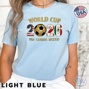 May include: Light blue t-shirt with "WORLD CUP 2026" in gold lettering. The numbers are designed with the flags of the USA, Canada, and Mexico. The text "USA CANADA MEXICO" is below the numbers. A "SHIP FROM USA" sticker is on the right.