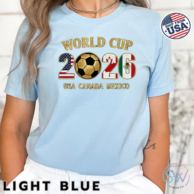 May include: Light blue t-shirt with "WORLD CUP 2026" in gold lettering. The numbers are designed with the flags of the USA, Canada, and Mexico. The text "USA CANADA MEXICO" is below the numbers. A "SHIP FROM USA" sticker is on the right.