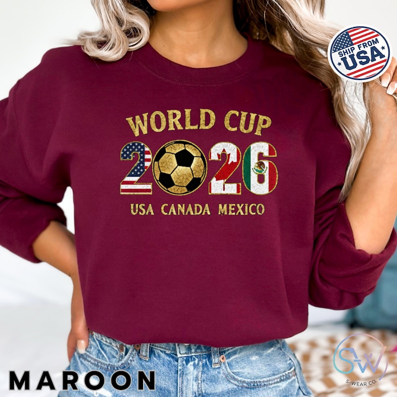 May include: Maroon sweatshirt with "WORLD CUP 2026" in gold lettering. The number 2 is designed with the American flag, and the 6 with the Mexican flag. A soccer ball is in the center. The text "USA CANADA MEXICO" is below the numbers.