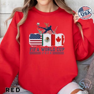 May include: Red sweatshirt featuring a FIFA World Cup 2026 graphic. The design includes flags of the USA, Mexico, and Canada, with a soccer player kicking a ball. The text "FIFA WORLD CUP 2026" is displayed below the flags.