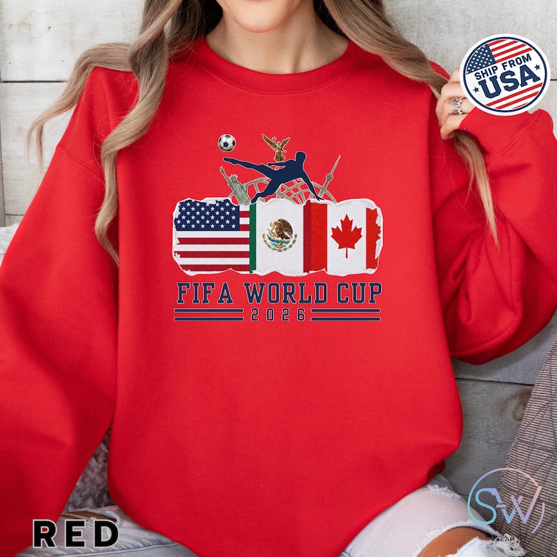 May include: Red sweatshirt featuring a FIFA World Cup 2026 graphic. The design includes flags of the USA, Mexico, and Canada, with a soccer player kicking a ball. The text "FIFA WORLD CUP 2026" is displayed below the flags.