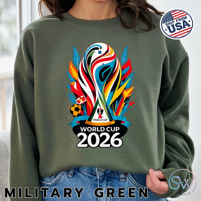 May include: An olive green crewneck sweatshirt featuring a colorful graphic of the World Cup 2026 trophy. The design includes abstract red, yellow, and blue shapes, a soccer ball, and the text "WORLD CUP 2026" printed in bold white letters.