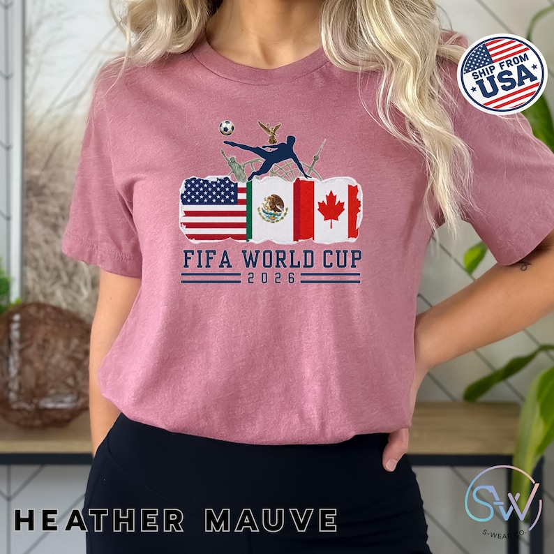 May include: Heather mauve t-shirt with a FIFA World Cup 2026 graphic. The design features flags of the USA, Mexico, and Canada, with a soccer player kicking a ball. A small USA flag logo is in the upper right corner.
