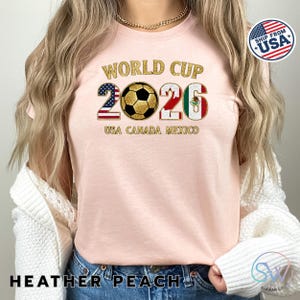 May include: Heather peach-colored t-shirt with "WORLD CUP 2026" in gold glitter lettering. The design includes a soccer ball and the flags of the USA, Canada, and Mexico. The text "USA CANADA MEXICO" is below the numbers.