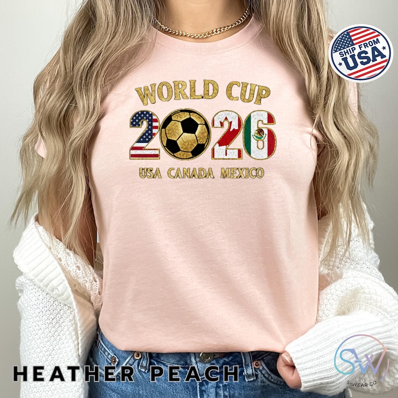 May include: Heather peach-colored t-shirt with "WORLD CUP 2026" in gold glitter lettering. The design includes a soccer ball and the flags of the USA, Canada, and Mexico. The text "USA CANADA MEXICO" is below the numbers.