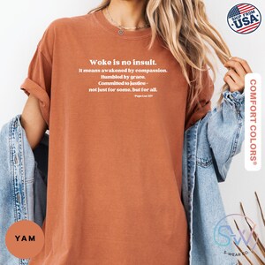 May include: A rust-colored t-shirt with the text "Woke is no insult" and a quote by Pope Leo XIV. The shirt is a Comfort Colors brand and has a relaxed fit. The image also includes a denim jacket.