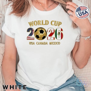 May include: White t-shirt with a gold-colored "World Cup 2026" graphic. The design includes a soccer ball and the flags of the USA, Canada, and Mexico. The text "USA CANADA MEXICO" is below the numbers.