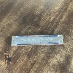 May include: A silver harmonica with the words "Melody Master" in cursive script. The harmonica has a rectangular shape with decorative edges and a red accent. The text "Made in Germany" is also visible.