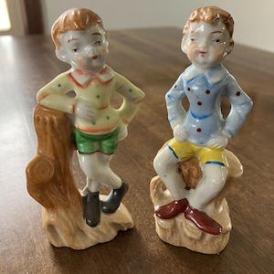 May include: Two vintage porcelain figurines of young boys. One boy wears a yellow shirt with green polka dots and green shorts, the other wears a blue shirt with red polka dots and yellow shorts. Both figurines have brown hair and are standing on a base.