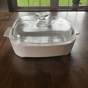May include: A white rectangular casserole dish with a clear glass lid. The dish has a clear glass knob on top of the lid and two white handles. The dish is sitting on a dark brown wooden table.