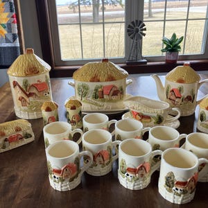 May include: A collection of ceramic kitchenware with a farmhouse theme. The set includes a canister, teapot, mugs, and other serving pieces. Each piece is decorated with a thatched roof design and a painted farm scene in shades of beige, red, and green.