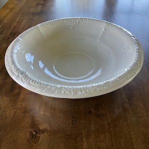 May include: A large, off-white ceramic bowl with a wide, decorative rim. The rim features a repeating pattern of raised, stylized floral and scrollwork designs. The bowl is set on a dark wooden surface.