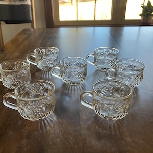 May include: A set of seven clear glass teacups with a detailed embossed grape and fruit design. Each cup has a small handle and sits on a wooden table. The cups are arranged in a circular pattern.