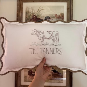 May include: A rectangular white throw pillow with a brown scalloped edge. The center features a dark brown line drawing of a cow standing in grass, with the text "THE TANNERS" printed in a simple, dark brown sans-serif font below the cow.