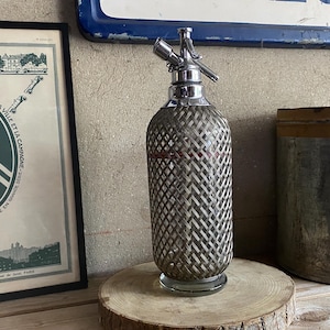 Könnte beinhalten: Vintage Soda-Siphon mit gewebtem Metall außen und Chrom-Oberteil. Die Glasflasche steht auf einer Holzscheibe. Im Hintergrund ist eine gerahmte Citroën-Werbung sowie ein Metallbehälter zu sehen.