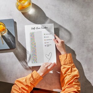 Peut inclure: Une personne tient une feuille "Self-Love Tracker" avec une grille et des cases &agrave; cocher. Un c&oelig;ur est dessin&eacute; en bas. Une chemise orange est visible. Un journal bleu et un verre d'eau avec des fruits sont sur la table.