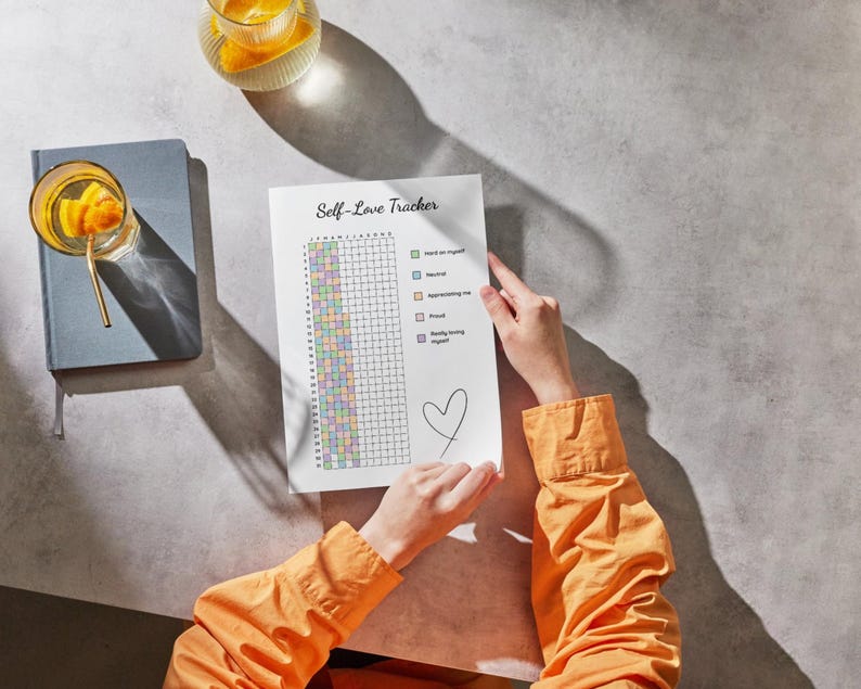 Peut inclure: Une personne tient une feuille "Self-Love Tracker" avec une grille et des cases &agrave; cocher. Un c&oelig;ur est dessin&eacute; en bas. Une chemise orange est visible. Un journal bleu et un verre d'eau avec des fruits sont sur la table.