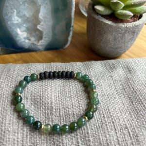 May include: A green beaded bracelet featuring black lava beads and gold-coloured spacers. The bracelet, made of small, round, green beads, rests on a grey, textured surface. A small succulent plant in a grey pot is in the background.