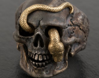May include: A detailed, dark silver skull ring with a gold-colored snake wrapped around it. The snake's head emerges from the eye socket, creating a striking contrast. The ring has a gothic or biker theme.
