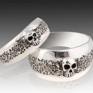 May include: Two silver skull rings with a textured design. The rings are stacked on top of each other.