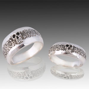 May include: Two silver skull rings with a textured band. The larger ring has a larger skull and the smaller ring has a smaller skull.