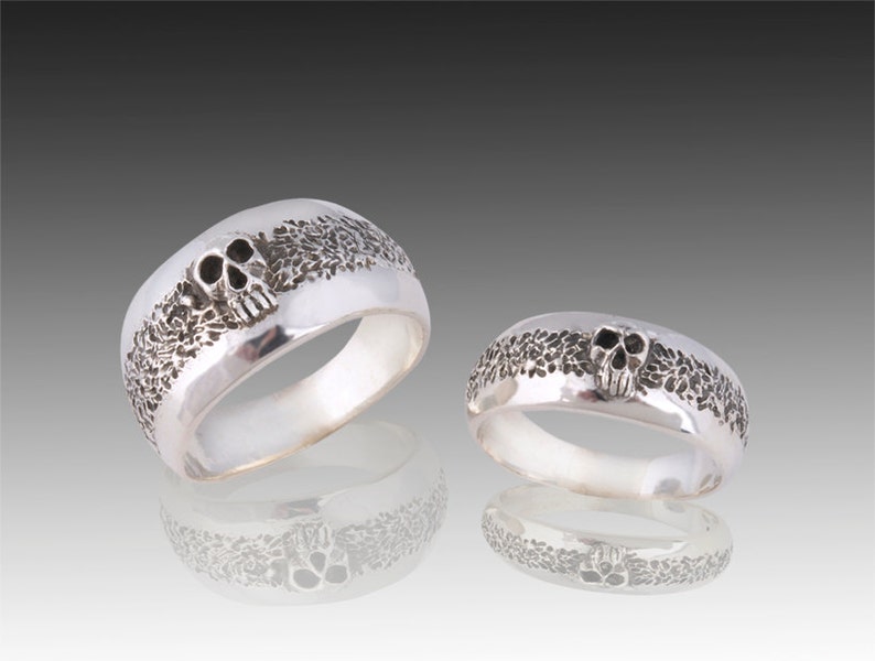 May include: Two silver skull rings with a textured band. The larger ring has a larger skull and the smaller ring has a smaller skull.
