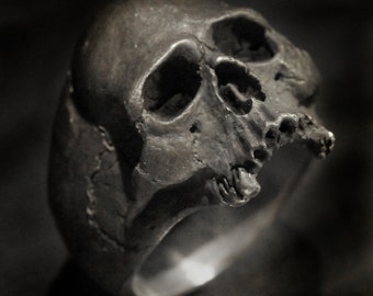 Könnte beinhalten: Ein silberner Totenkopfring mit einem detaillierten Design. Der Ring ist aus Metall und hat eine strukturierte Oberfläche.