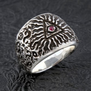 May include: A silver ring with a textured surface and a red gemstone in the center of a triangle design. The triangle is surrounded by a sunburst pattern.
