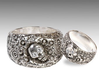 May include: Two silver rings with a skull design. The skull has two clear gemstones for eyes. The rings are textured with a swirling pattern.