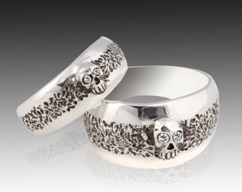 May include: Two silver skull rings with diamond eyes. The rings are textured and have a detailed skull design.