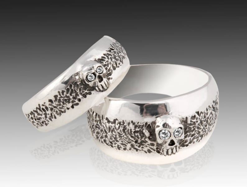 May include: Two silver skull rings with diamond eyes. The rings are textured and have a detailed skull design.