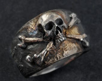 Könnte beinhalten: Ein silberner Ring mit Totenkopf und gekreuzten Knochen, mit einer strukturierten Oberfläche. Der Totenkopf ist detailliert und die gekreuzten Knochen sind ineinander verschlungen.
