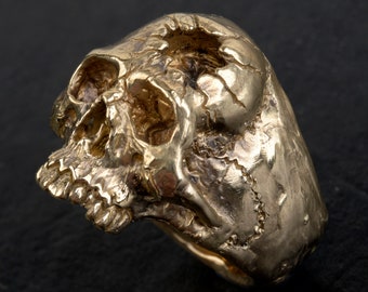 Könnte beinhalten: Ein goldener Totenkopfring mit einem detaillierten, strukturierten Design. Der Ring ist aus Metall und hat eine Totenkopfform mit einem markanten Unterkiefer.