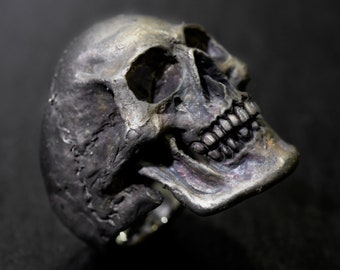 Könnte beinhalten: Ein silberner Totenkopfring mit einem detaillierten Design. Der Totenkopf ist nach rechts gerichtet und hat einen leicht geöffneten Mund mit sichtbaren Zähnen.
