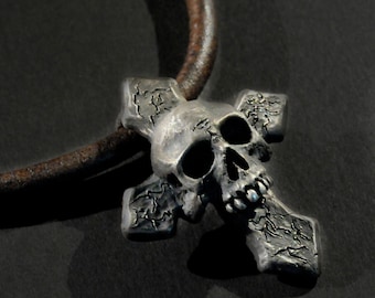 May include: A silver skull pendant with a cross design. The pendant is on a brown leather cord.