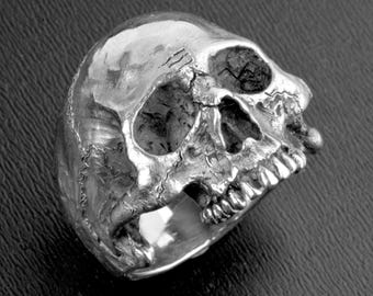 May include: A silver skull ring with detailed features, including teeth and eye sockets.