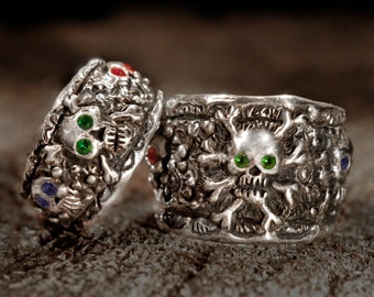 May include: Two silver skull rings with green and red gemstones. The rings are detailed with intricate carvings and are a gothic style.